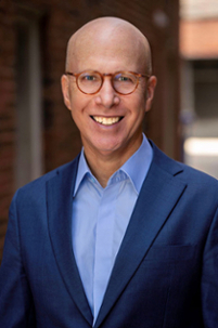 jonathan eig wearing eye glasses smiling at the camera in a light blue shirt and dark blue blazer