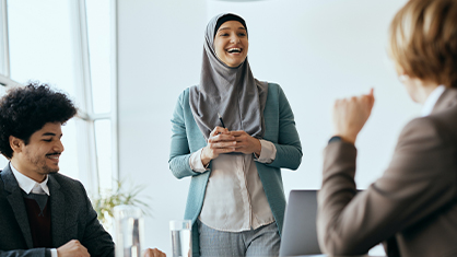 A woman in a hijab stands and smiles while speaking to two seated colleagues in a modern office setting.